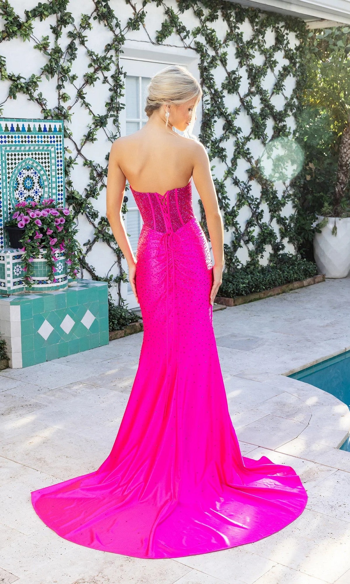 A woman in a bright pink, strapless Portia and Scarlett XO098 long prom dress stands by a pool with her back to the camera, surrounded by lush greenery and decorative tiles.